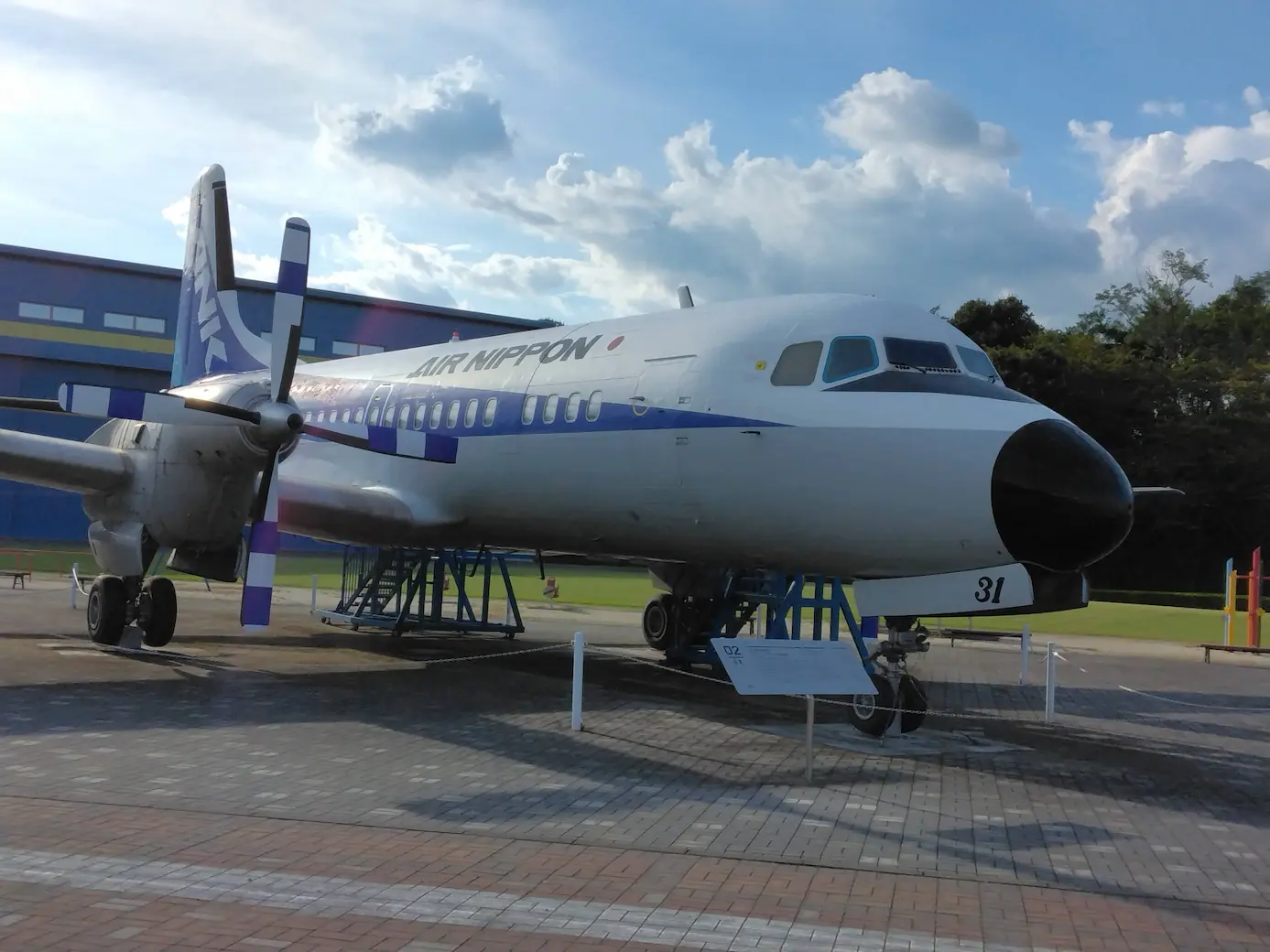 野外に展示された飛行機の写真。奥には、青い建物がある。上半分は、晴れた空だが、画面上部右側の方に固まった雲がのびている